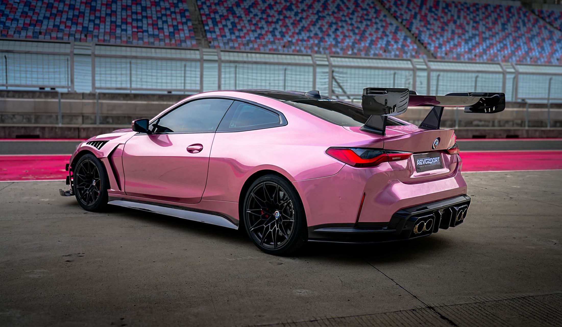 Pink BMW G82 M4 fitted with a REVOZPORT carbon fiber widebody aero kit, front splitter, hood vents, and side skirts on a race track pit lane.
