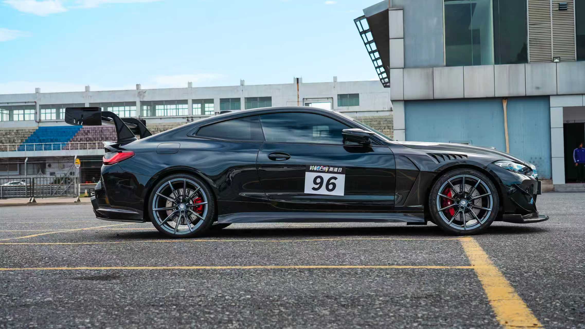 Black BMW G82 M4 track build with carbon fiber vented fenders, side skirts, rear diffuser, and oversized GT wing parked at a professional racing circuit pit lane.