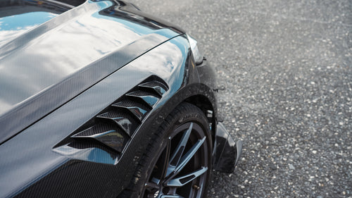 Black BMW G82 M4 track build with carbon fiber vented fenders, side skirts, rear diffuser, and oversized GT wing parked at a professional racing circuit pit lane.