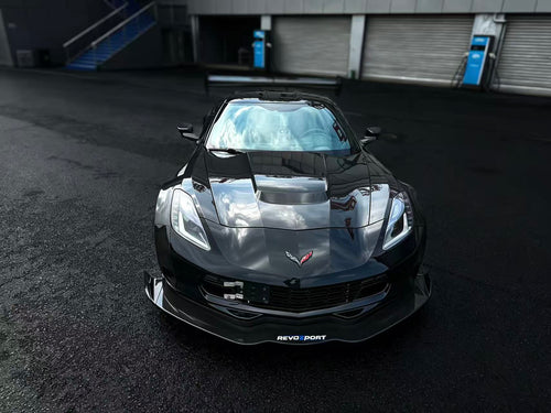 Black Chevrolet Corvette race car parked on a wet industrial track surface, featuring an aggressive wide-body kit, large rear wing, carbon fiber aerodynamic components, and lightweight performance wheels with red brake calipers. The moody urban backdrop and reflective pavement emphasize the car’s high-performance motorsport styling.