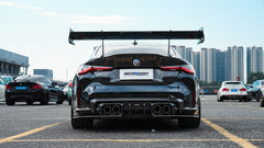 Black BMW G82 M4 track build with carbon fiber vented fenders, side skirts, rear diffuser, and oversized GT wing parked at a professional racing circuit pit lane.