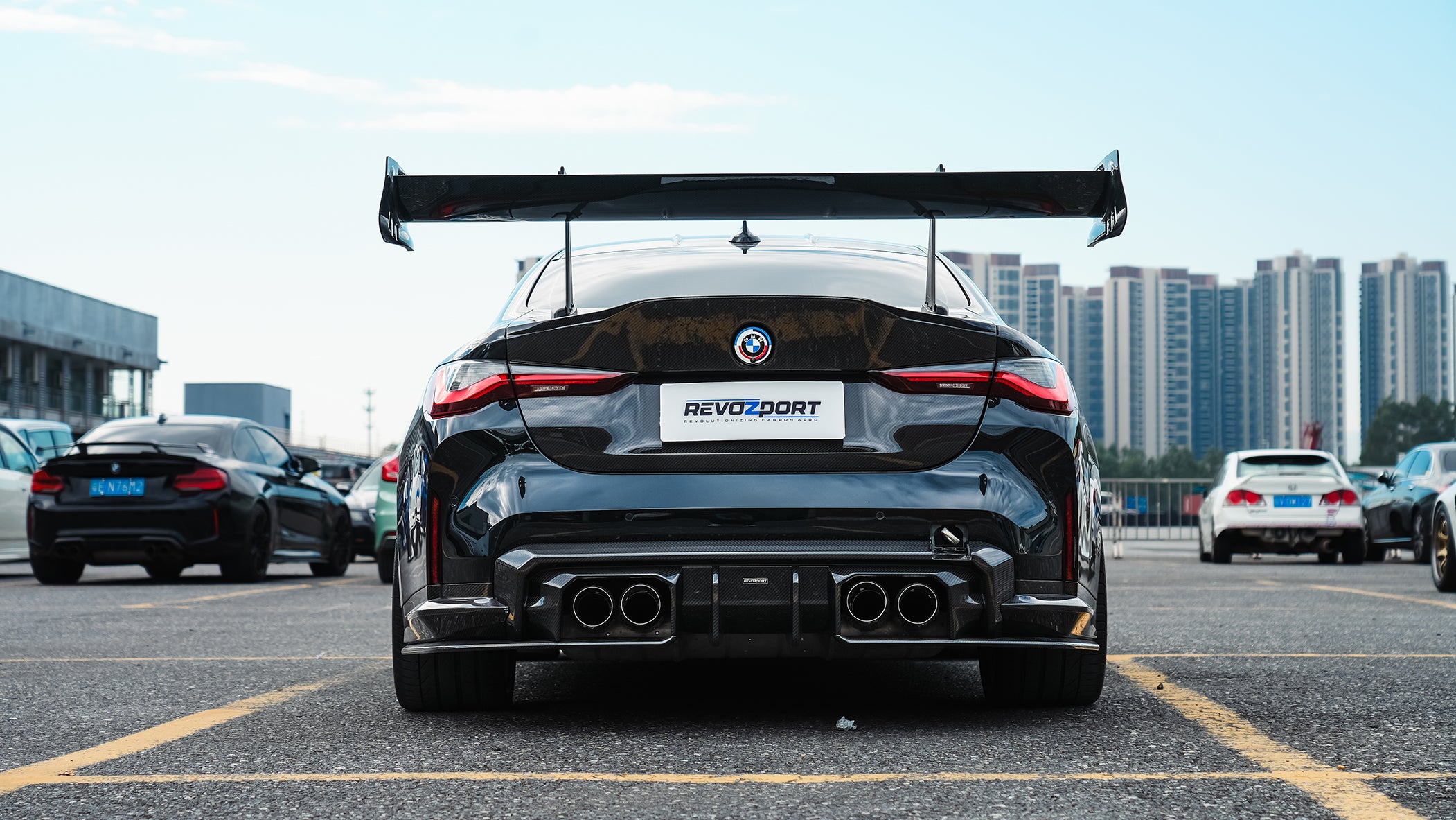 Black BMW G82 M4 track build with carbon fiber vented fenders, side skirts, rear diffuser, and oversized GT wing parked at a professional racing circuit pit lane.