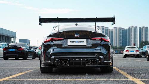 Black BMW G82 M4 track build with carbon fiber vented fenders, side skirts, rear diffuser, and oversized GT wing parked at a professional racing circuit pit lane.