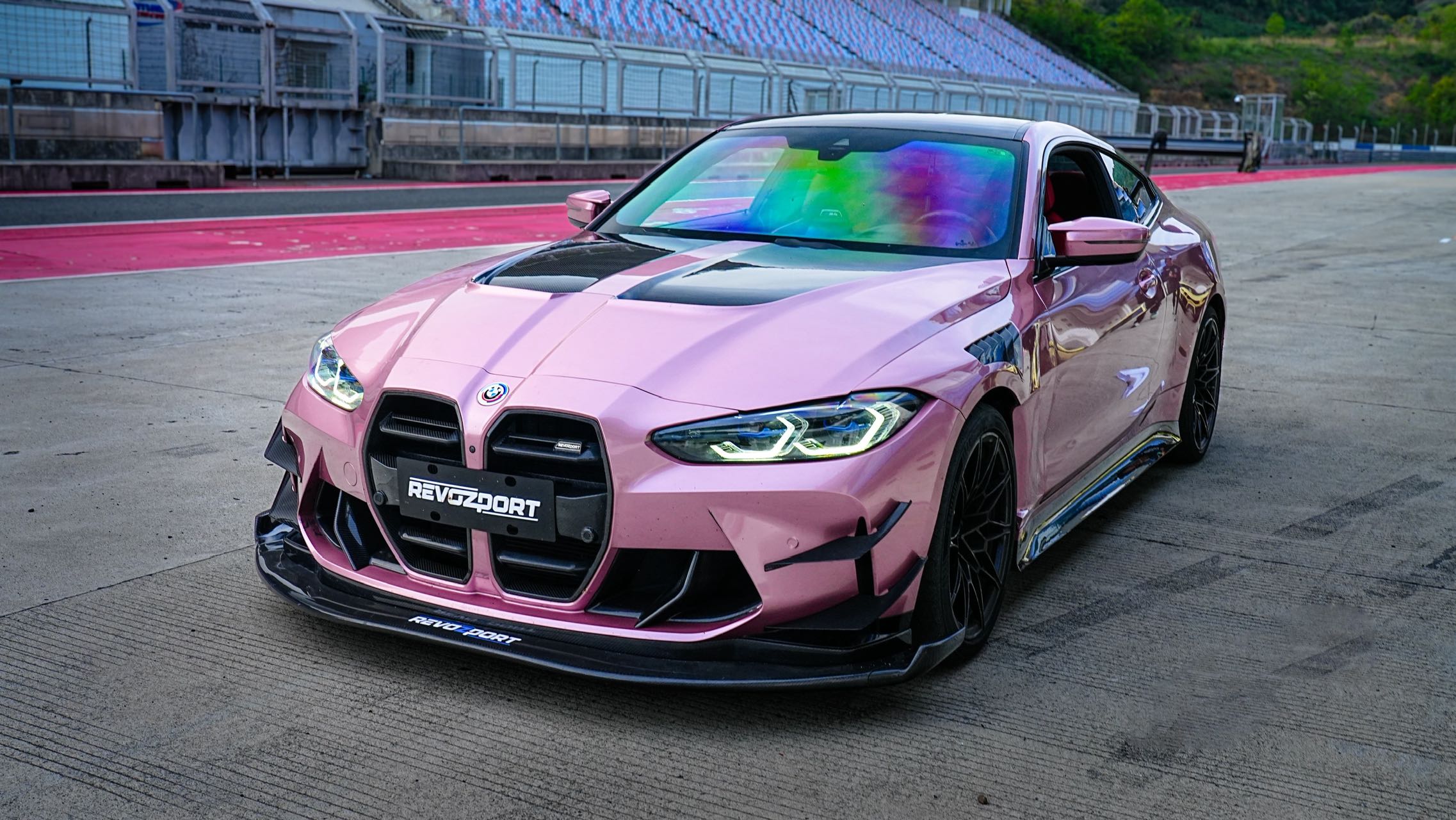 Pink BMW G82 M4 fitted with a REVOZPORT carbon fiber widebody aero kit, front splitter, hood vents, and side skirts on a race track pit lane.
