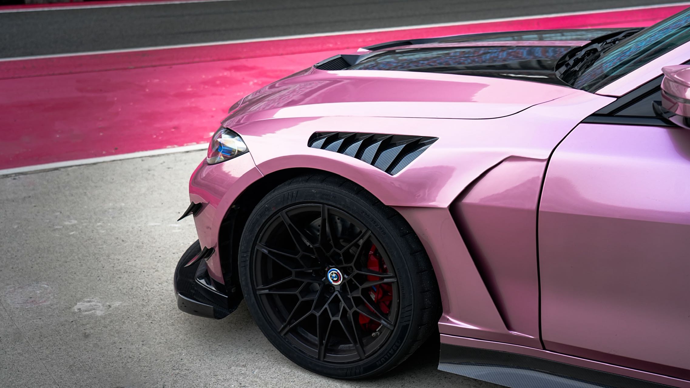 Pink BMW G82 M4 fitted with a REVOZPORT carbon fiber widebody aero kit, front splitter, hood vents, and side skirts on a race track pit lane.