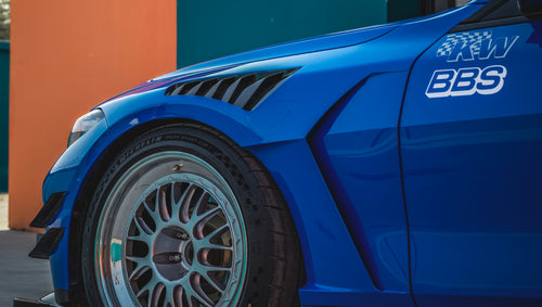 Blue BMW G82 M4 with carbon fiber front splitter, side skirts, rear wing, and aftermarket wheels parked in a modern industrial courtyard with colorful architectural walls.
