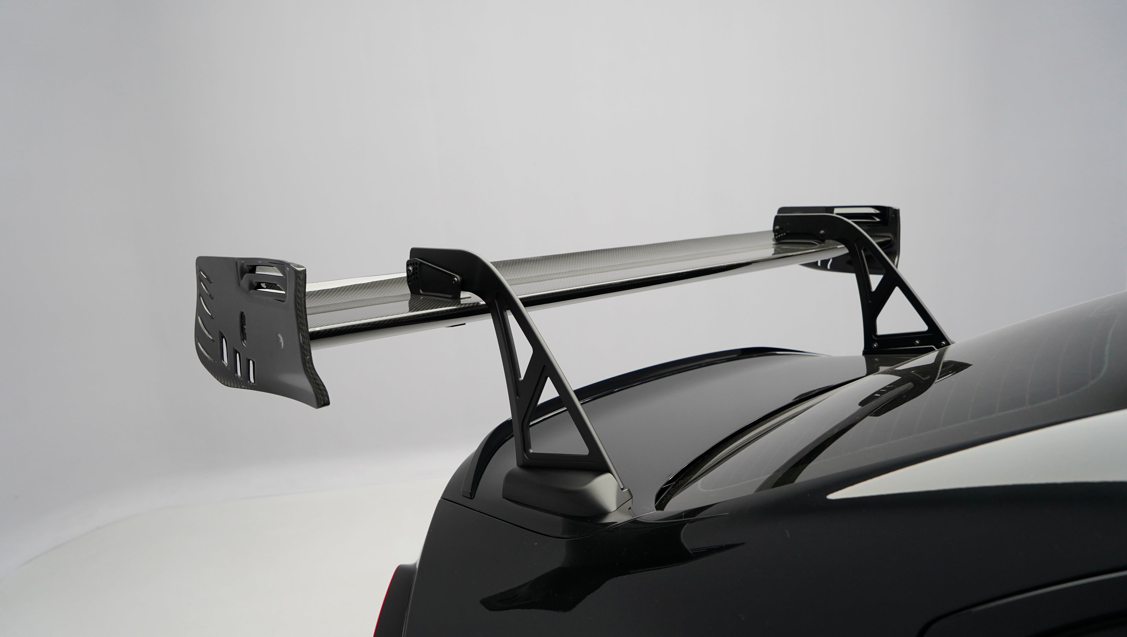 Front close-up of a black BMW G87 M2 featuring an aggressive carbon fiber front bumper, deep front splitter, large air intakes, and gloss carbon fiber aero details in a studio setting.
