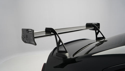 Front close-up of a black BMW G87 M2 featuring an aggressive carbon fiber front bumper, deep front splitter, large air intakes, and gloss carbon fiber aero details in a studio setting.
