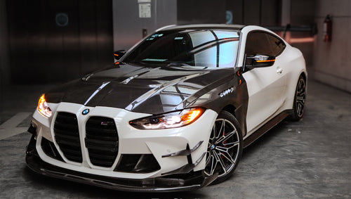 White BMW G82 M4 featuring a forged carbon fiber hood, front splitter, side canards, and aggressive aero styling parked inside a modern underground garage.