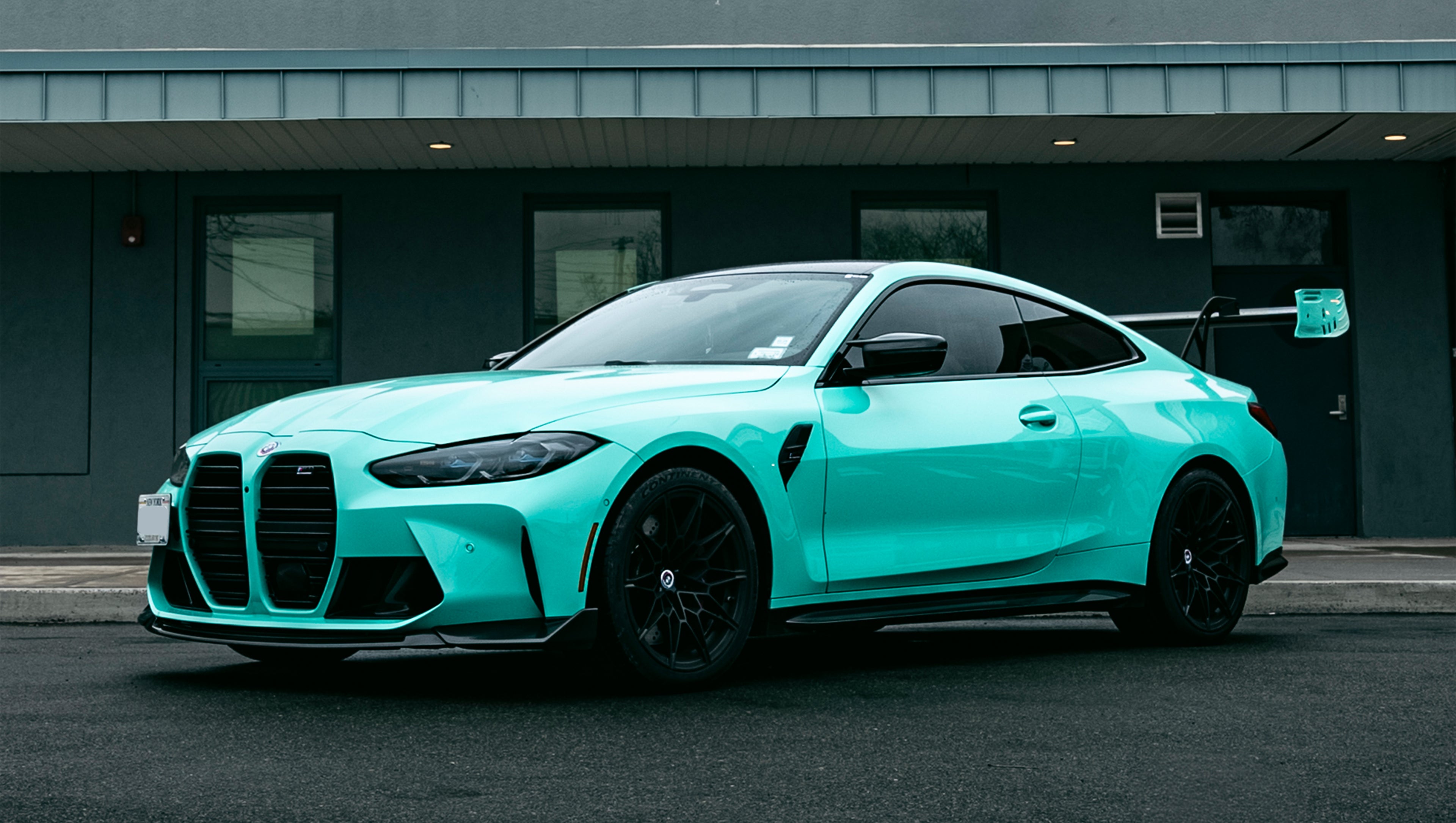 Mint green BMW G82 M4 with carbon fiber front splitter, side skirts, and large rear wing parked on a wet urban street outside a modern building.