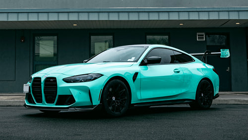 Mint green BMW G82 M4 with carbon fiber front splitter, side skirts, and large rear wing parked on a wet urban street outside a modern building.