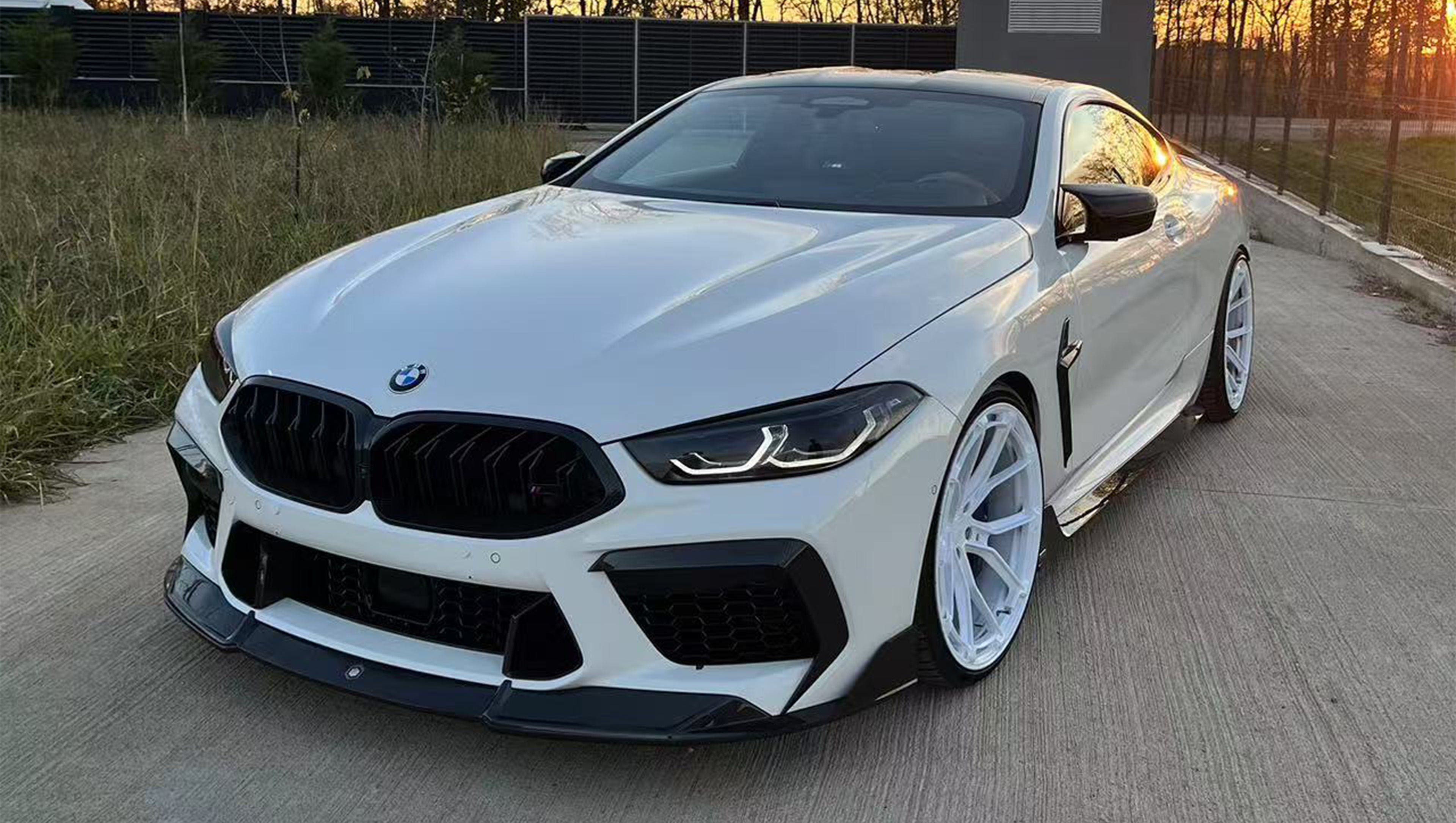 White BMW F92 M8 featuring a REVOZPORT carbon fiber front lip, side skirts, black front grille, and white multi-spoke forged wheels parked at sunset on an open road.