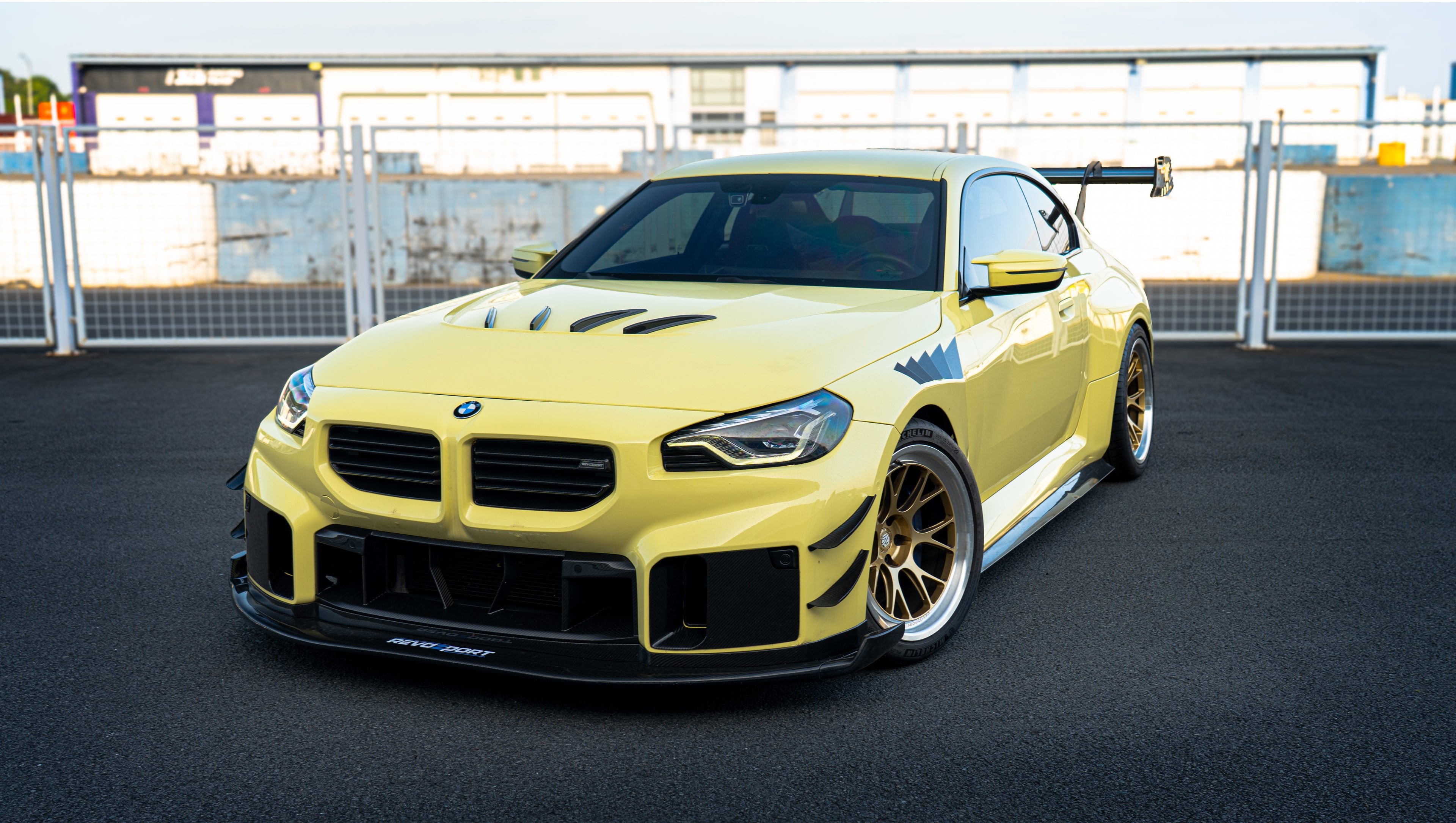 Pastel yellow BMW G82 M4 with carbon fiber front splitter, hood vents, side canards, bronze wheels, and large rear wing parked at a race track facility during golden hour.