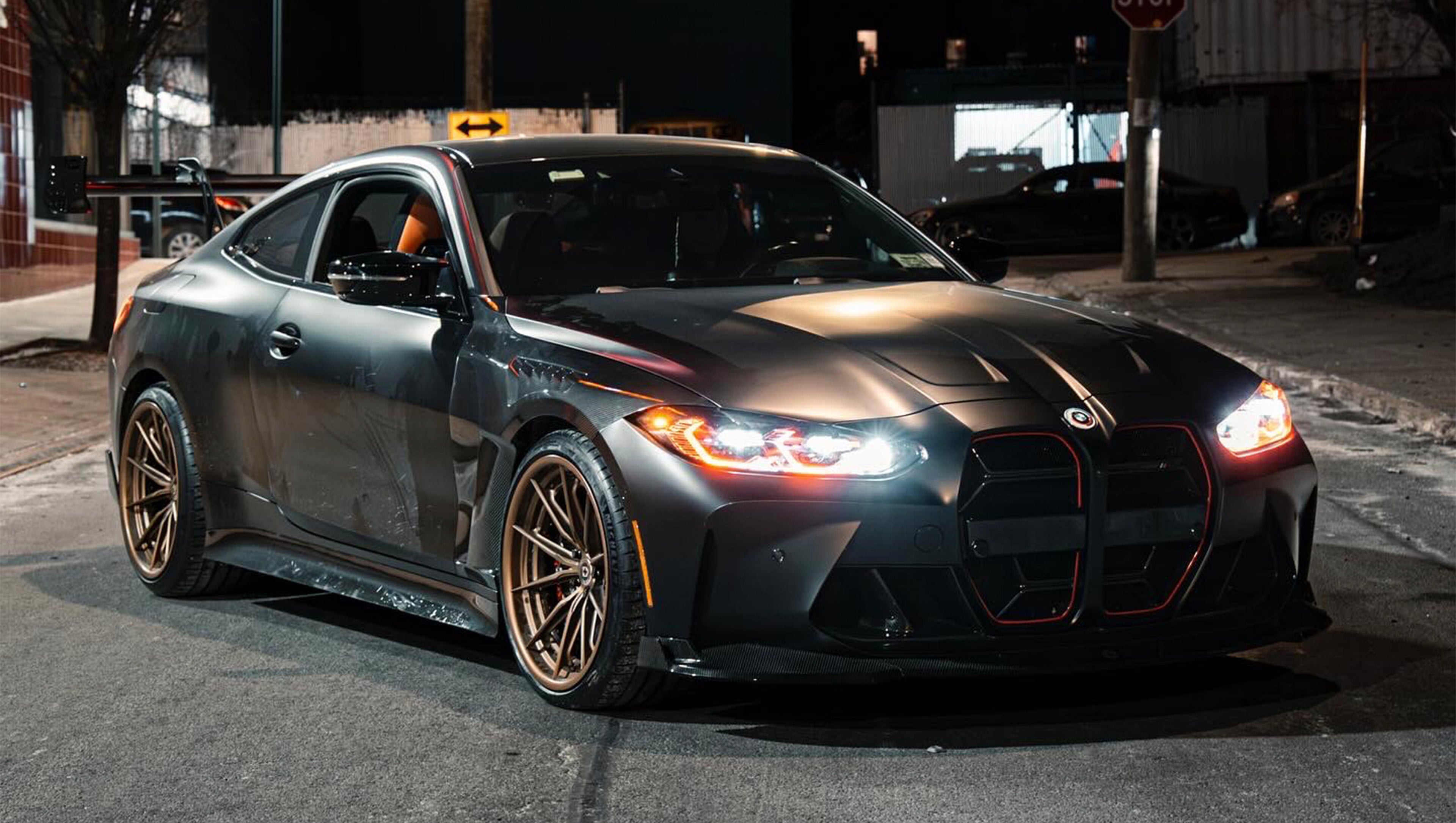Matte black BMW G82 M4 with carbon fiber front splitter, side skirts, vented hood, bronze wheels, and oversized rear wing parked on a city street at night with headlights on.
