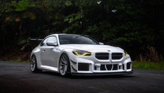 White widebody BMW G82 M4 with aggressive front bumper, canards, deep front splitter, large rear wing, and yellow LED headlights parked on a wet road surrounded by dense tropical forest.