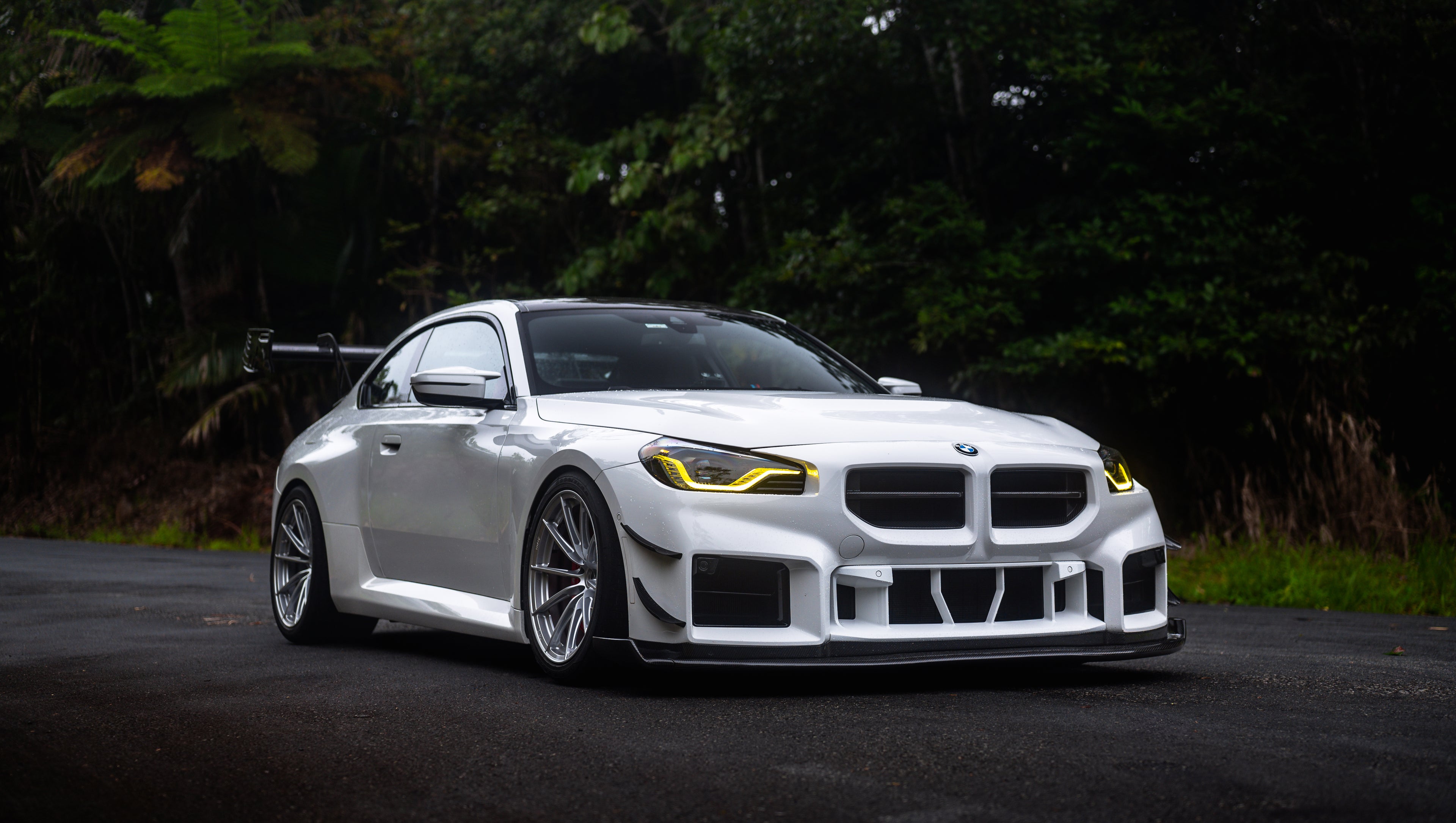 White widebody BMW G82 M4 with aggressive front bumper, canards, deep front splitter, large rear wing, and yellow LED headlights parked on a wet road surrounded by dense tropical forest.