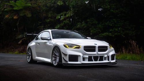White widebody BMW G82 M4 with aggressive front bumper, canards, deep front splitter, large rear wing, and yellow LED headlights parked on a wet road surrounded by dense tropical forest.