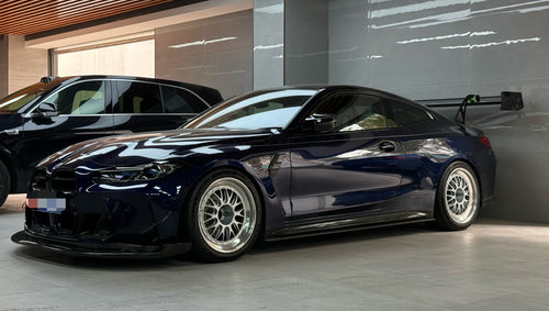Dark blue BMW G82 M4 equipped with carbon fiber front splitter, side skirts, vented fenders, and a large rear wing, parked inside a modern luxury garage.
