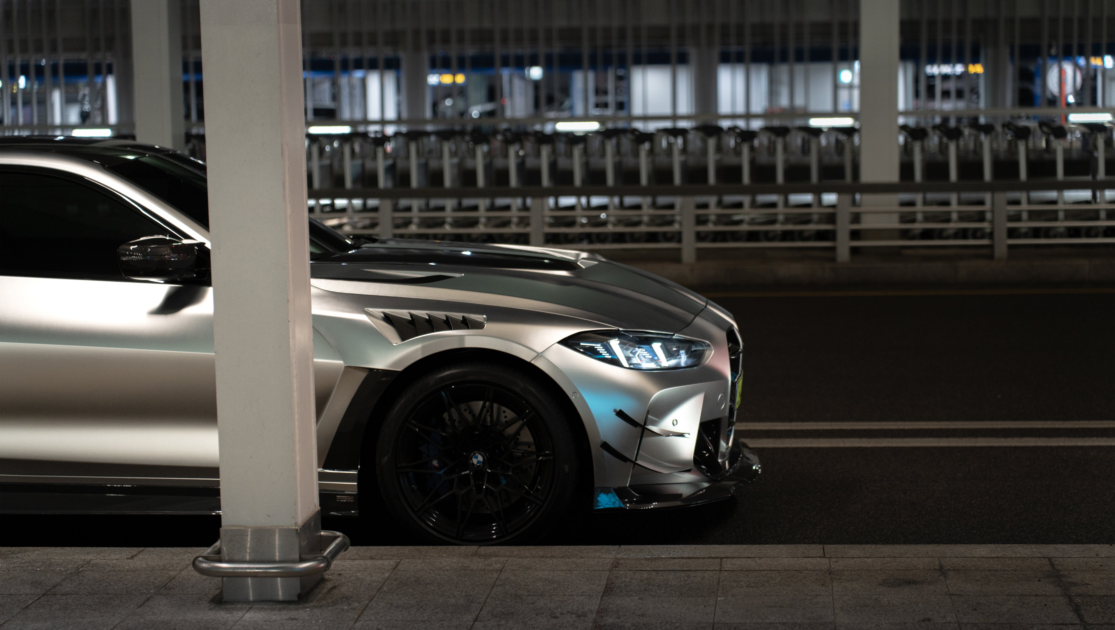Matte gray BMW G82 M4 featuring an aggressive REVOZPORT carbon fiber aero kit with vented hood, front canards, wide fender vents, and illuminated LED headlights in a nighttime parking garage.