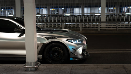 Matte gray BMW G82 M4 featuring an aggressive REVOZPORT carbon fiber aero kit with vented hood, front canards, wide fender vents, and illuminated LED headlights in a nighttime parking garage.