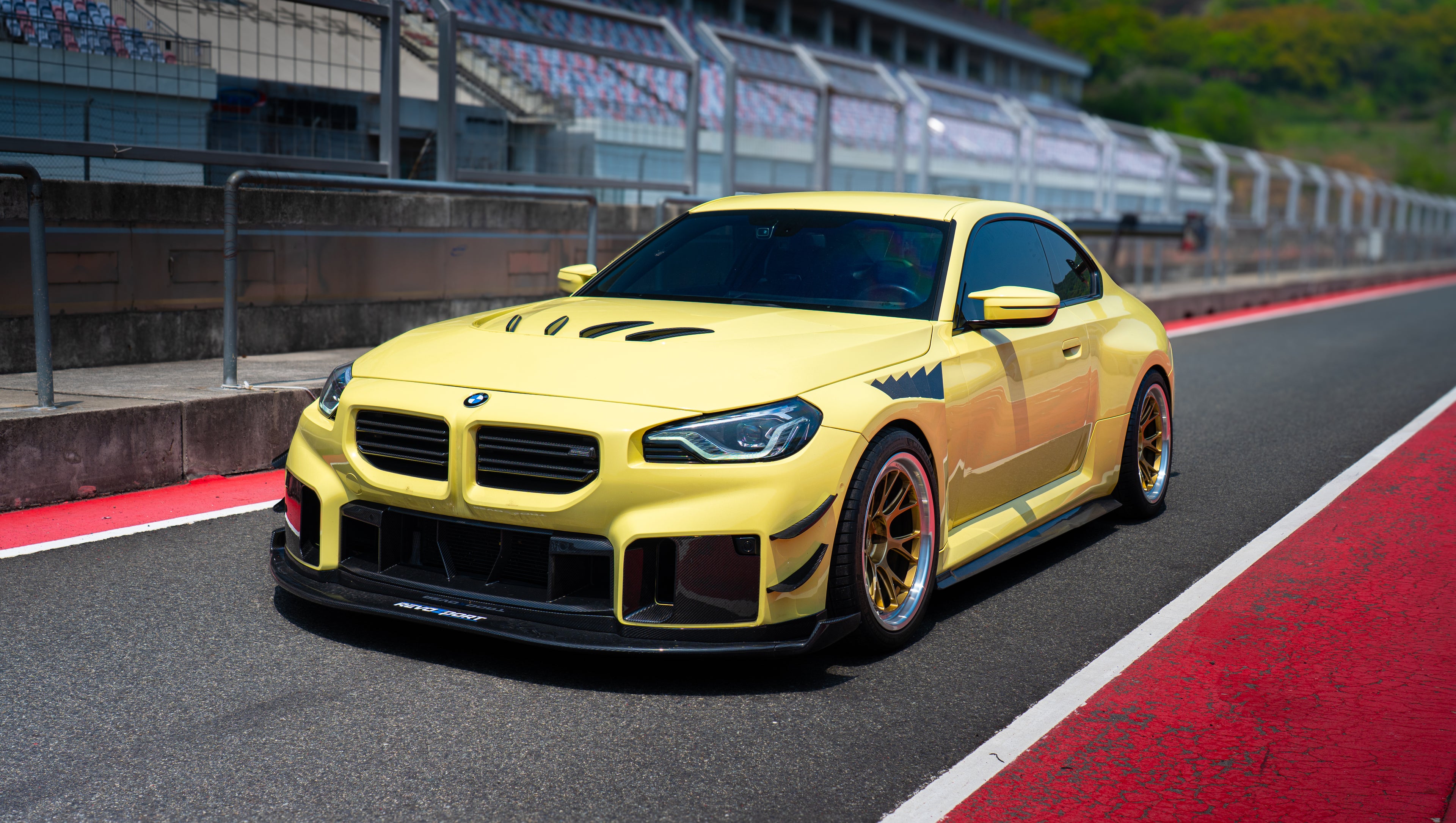 Yellow BMW G82 M4 with aggressive carbon fiber front splitter, vented hood, side canards, and bronze wheels parked on a race track pit lane.