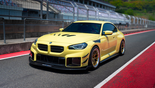 Yellow BMW G82 M4 with aggressive carbon fiber front splitter, vented hood, side canards, and bronze wheels parked on a race track pit lane.