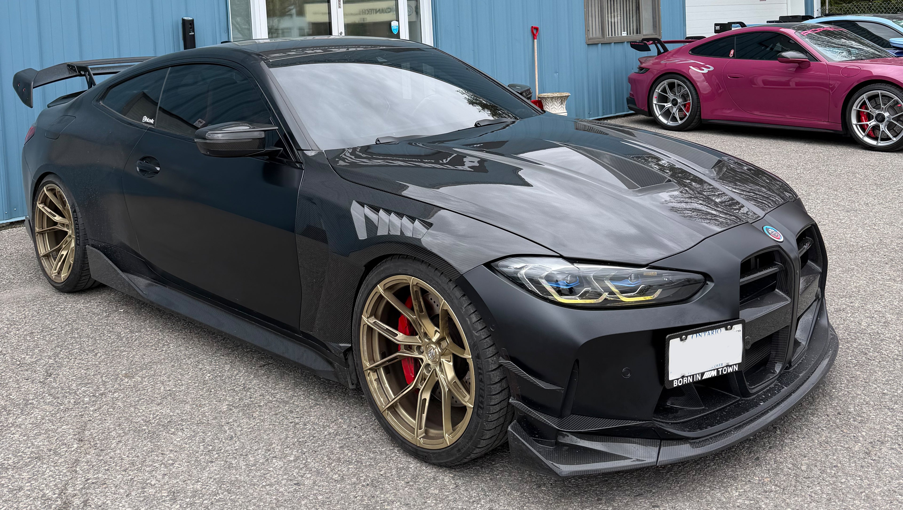 Matte black BMW G82 M4 featuring a REVOZPORT carbon fiber body kit with vented hood, front lip, side skirts, rear wing, and bronze forged wheels parked outside a workshop.