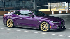 Purple BMW G82 M4 featuring a REVOZPORT carbon fiber body kit with front lip, side skirts, vented fenders, and gold wheels parked outside a modern industrial building.