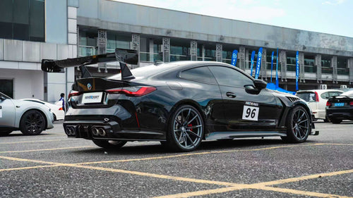Black BMW G82 M4 track build with carbon fiber vented fenders, side skirts, rear diffuser, and oversized GT wing parked at a professional racing circuit pit lane.