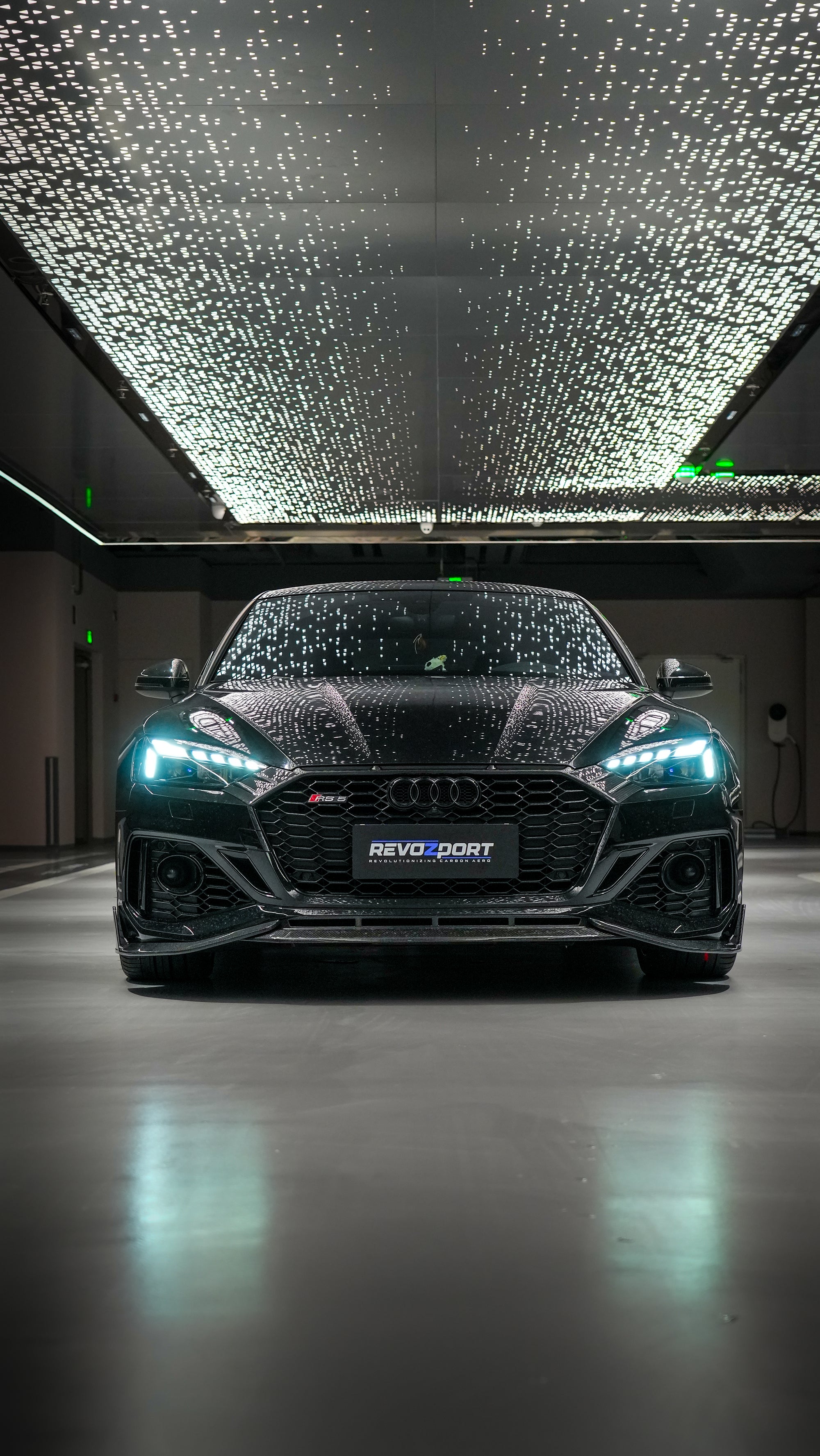Front view of a black Audi RS5 Sportback featuring a REVOZPORT carbon fiber body kit with aggressive front lip, side aero details, and illuminated LED headlights inside a modern luxury garage.