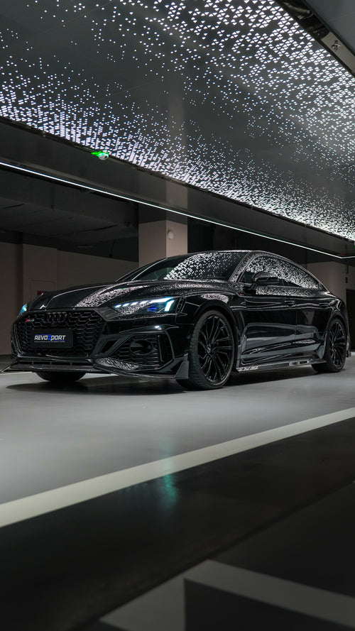 Front view of a black Audi RS5 Sportback featuring a REVOZPORT carbon fiber body kit with aggressive front lip, side aero details, and illuminated LED headlights inside a modern luxury garage.