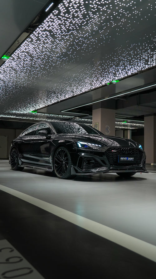 Front view of a black Audi RS5 Sportback featuring a REVOZPORT carbon fiber body kit with aggressive front lip, side aero details, and illuminated LED headlights inside a modern luxury garage.