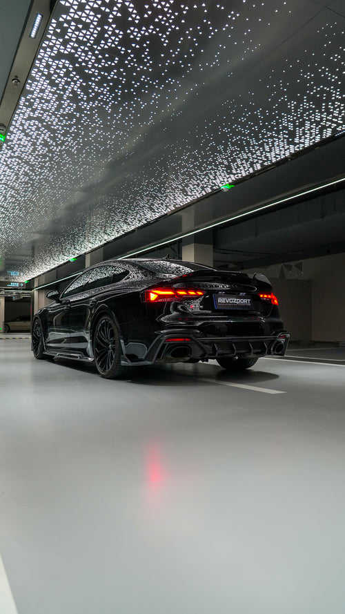 Front view of a black Audi RS5 Sportback featuring a REVOZPORT carbon fiber body kit with aggressive front lip, side aero details, and illuminated LED headlights inside a modern luxury garage.