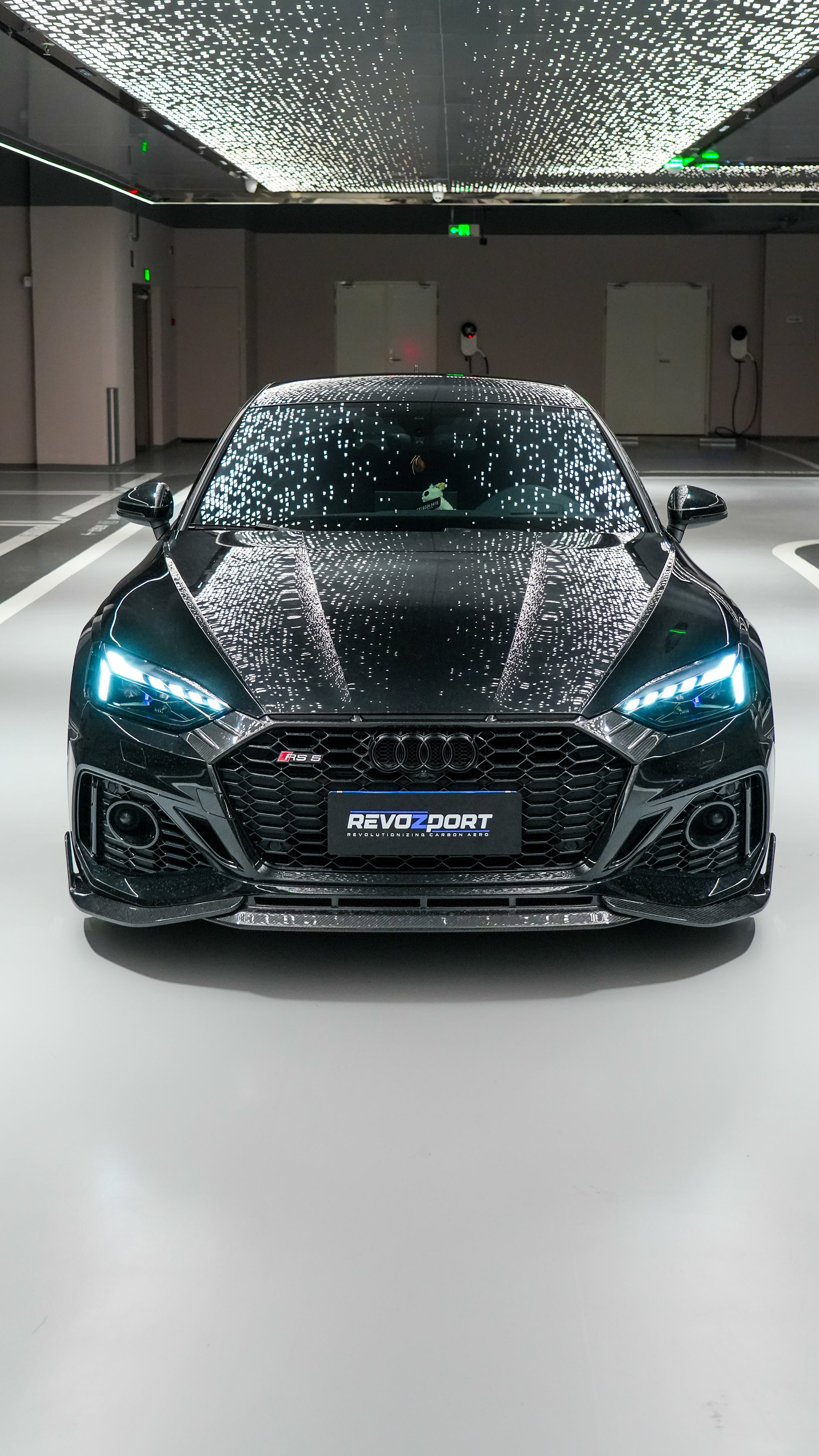 Front view of a black Audi RS5 Sportback featuring a REVOZPORT carbon fiber body kit with aggressive front lip, side aero details, and illuminated LED headlights inside a modern luxury garage.