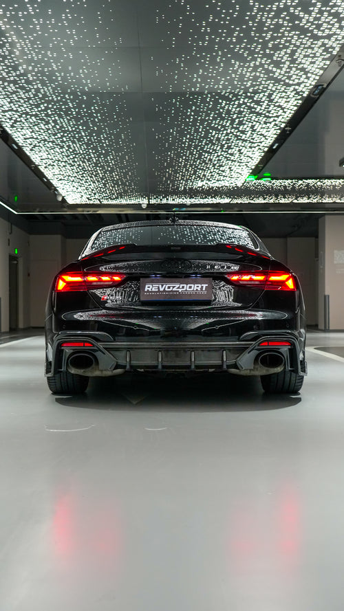 Front view of a black Audi RS5 Sportback featuring a REVOZPORT carbon fiber body kit with aggressive front lip, side aero details, and illuminated LED headlights inside a modern luxury garage.