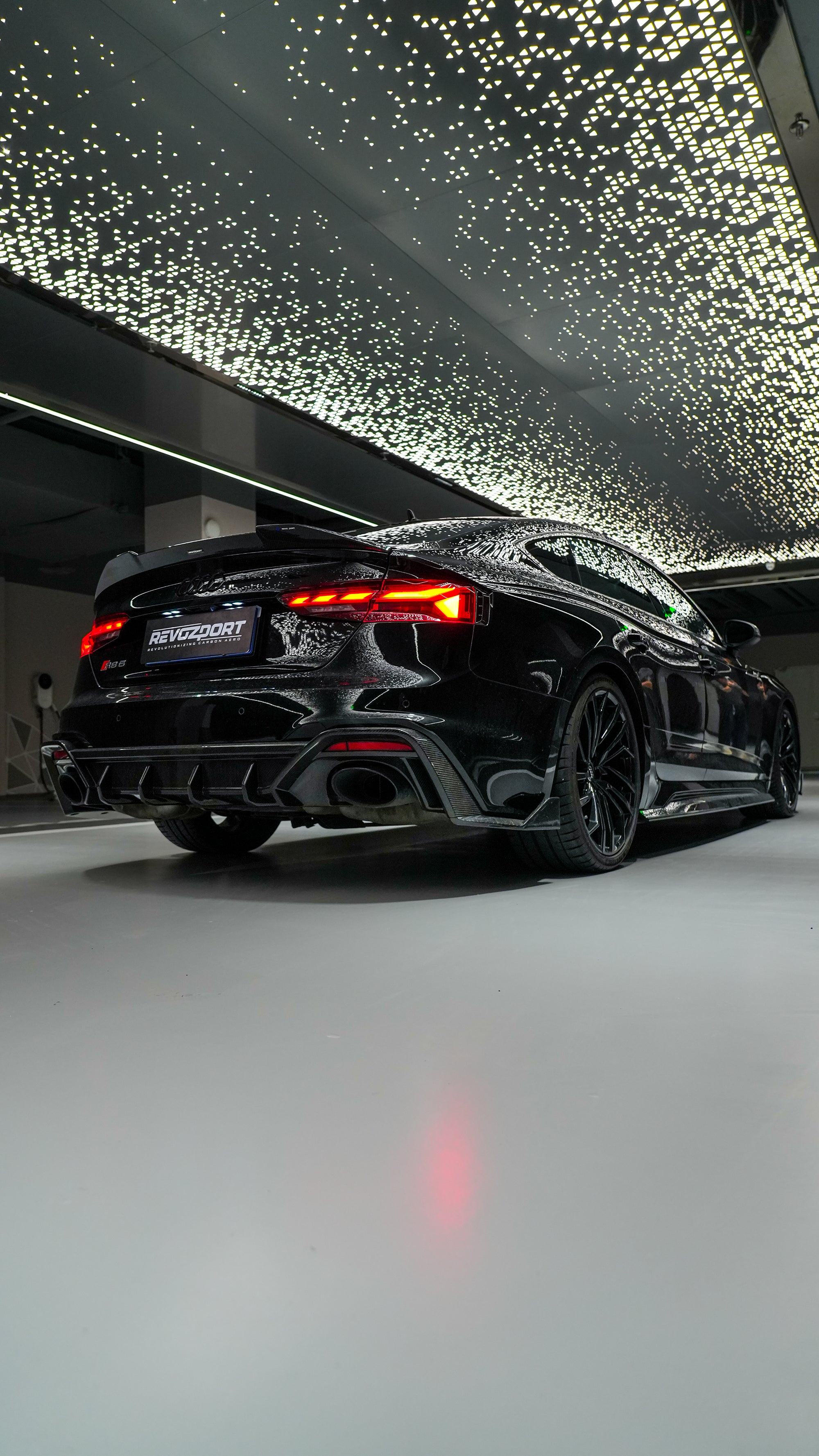Front view of a black Audi RS5 Sportback featuring a REVOZPORT carbon fiber body kit with aggressive front lip, side aero details, and illuminated LED headlights inside a modern luxury garage.