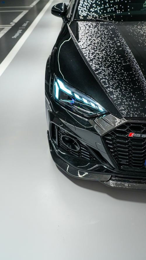 Front view of a black Audi RS5 Sportback featuring a REVOZPORT carbon fiber body kit with aggressive front lip, side aero details, and illuminated LED headlights inside a modern luxury garage.