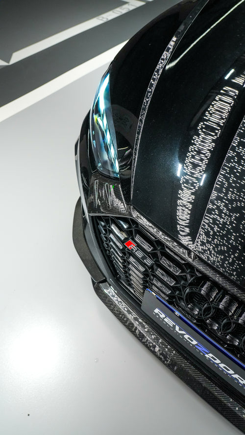 Front view of a black Audi RS5 Sportback featuring a REVOZPORT carbon fiber body kit with aggressive front lip, side aero details, and illuminated LED headlights inside a modern luxury garage.