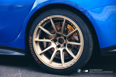 Blue BMW G80 M3 with REVOZPORT carbon fiber front splitter, side skirts, vented fenders, and large rear wing parked in a landscaped outdoor plaza.