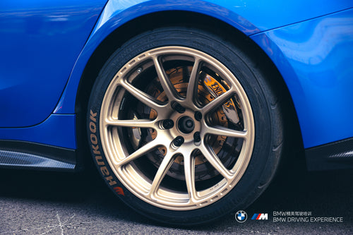 Blue BMW G80 M3 with REVOZPORT carbon fiber front splitter, side skirts, vented fenders, and large rear wing parked in a landscaped outdoor plaza.