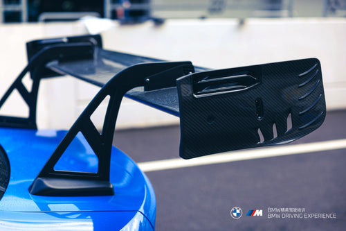 Blue BMW G80 M3 with REVOZPORT carbon fiber front splitter, side skirts, vented fenders, and large rear wing parked in a landscaped outdoor plaza.