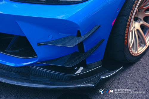 Blue BMW G80 M3 with REVOZPORT carbon fiber front splitter, side skirts, vented fenders, and large rear wing parked in a landscaped outdoor plaza.
