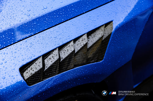 Blue BMW G80 M3 with REVOZPORT carbon fiber front splitter, side skirts, vented fenders, and large rear wing parked in a landscaped outdoor plaza.