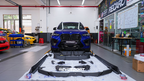 Blue BMW X5M featuring an aggressive REVOZPORT carbon fiber body kit with front lip, side skirts, enlarged front air intakes, and sporty aero styling displayed inside a modern performance workshop.
