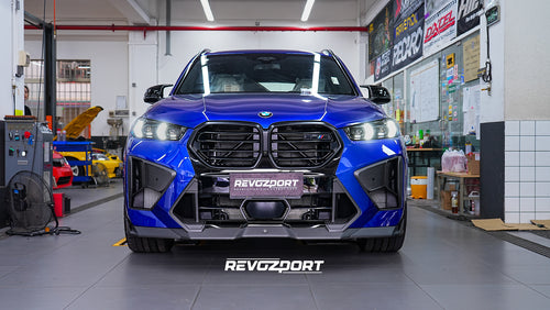 Blue BMW X5M featuring an aggressive REVOZPORT carbon fiber body kit with front lip, side skirts, enlarged front air intakes, and sporty aero styling displayed inside a modern performance workshop.
