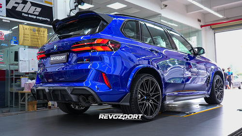 Blue BMW X5M featuring an aggressive REVOZPORT carbon fiber body kit with front lip, side skirts, enlarged front air intakes, and sporty aero styling displayed inside a modern performance workshop.