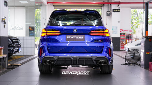 Blue BMW X5M featuring an aggressive REVOZPORT carbon fiber body kit with front lip, side skirts, enlarged front air intakes, and sporty aero styling displayed inside a modern performance workshop.