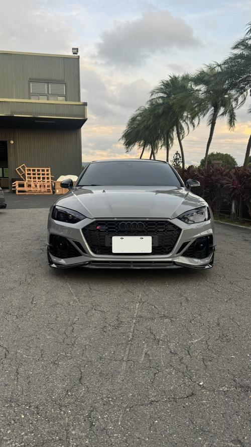 Gray Audi RS5 Sportback featuring a REVOZPORT carbon fiber front lip, side skirts, and aggressive front aero styling parked outdoors near tropical landscaping.