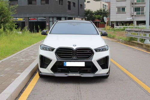 Front view of a white BMW X6M featuring an aggressive REVOZPORT front bumper and carbon fiber aero styling parked on a quiet urban street.