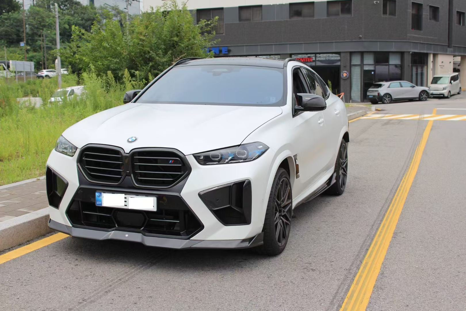 Front view of a white BMW X6M featuring an aggressive REVOZPORT front bumper and carbon fiber aero styling parked on a quiet urban street.